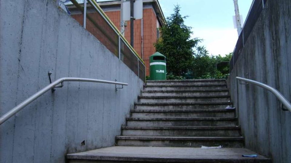 Scalinata in cemento che sale tra due muri alti, con corrimano metallici su entrambi i lati. In cima si vedono un edificio in mattoni, un cassonetto verde e alcuni alberi sotto un cielo parzialmente nuvoloso.