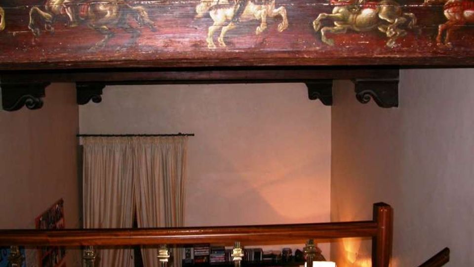 Interno di una casa con soppalco in legno e parapetto con colonnine, illuminato da una lampada da terra. In alto si vede una grande trave decorata con un dipinto di cavalieri, e sul fondo una tenda e scaffali con libri.