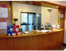 Banco reception in legno con ampia vetrata e sportello di servizio, con opuscoli e documenti esposti sul piano. Sullo sfondo si vede un corridoio con porta vetrata e pareti chiare illuminate da faretti a soffitto.
