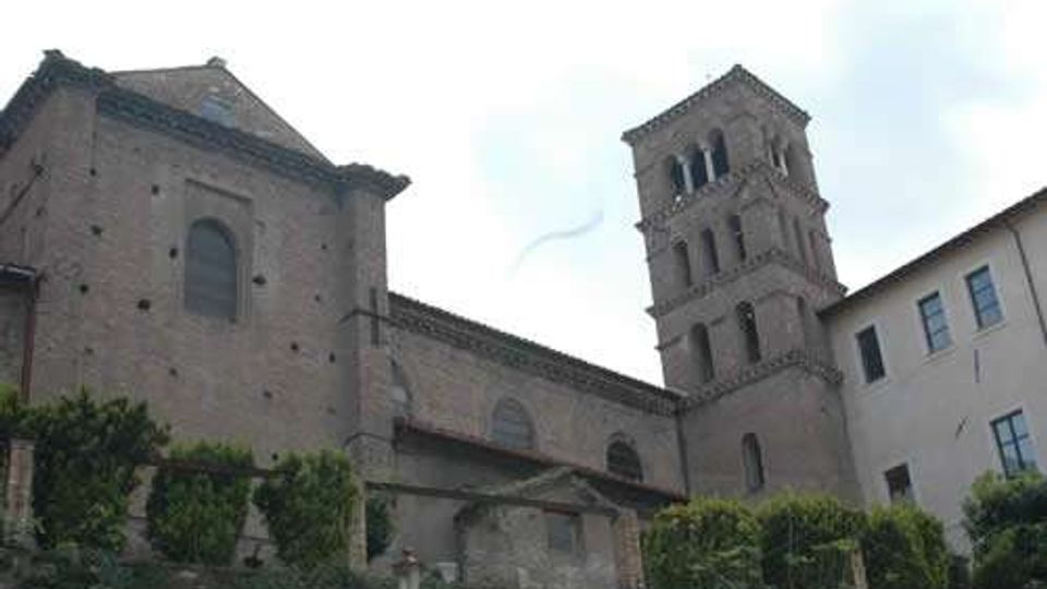Chiesa in muratura con facciate in mattoni e tetti in coppi, affiancata da un campanile quadrato a più livelli con finestre ad arco. L’edificio si affaccia su un pendio con vegetazione e arbusti, con un altro fabbricato adiacente sulla destra.