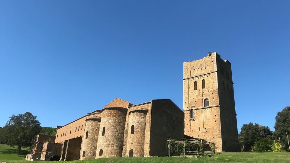 Edificio storico in pietra con torri cilindriche e un’alta torre quadrata, situato su un prato verde. La struttura presenta piccole finestre ad arco ed è circondata da alberi sotto un cielo sereno.