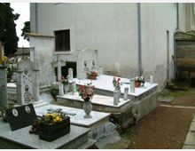 Piccolo cimitero con tombe in marmo e lapidi con croci e vasi di fiori, disposte su piattaforme rialzate. Sullo sfondo si vede il muro esterno chiaro di un edificio e un vialetto laterale in cemento.