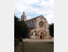 Facciata di una chiesa storica in pietra chiara con grande rosone circolare e portale ad arco, affacciata su una piazza in pavé. Sul lato sinistro si vede il campanile e intorno alberi e colline sullo sfondo.