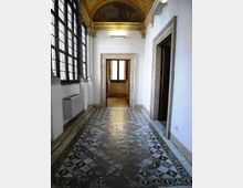 Corridoio interno di un edificio storico con pavimento in piastrelle decorative a motivi geometrici, grandi finestre a griglia sul lato sinistro e portali in pietra. Il soffitto è a volta con affreschi e cornici dorate, con un passaggio che conduce a una stanza illuminata in fondo.