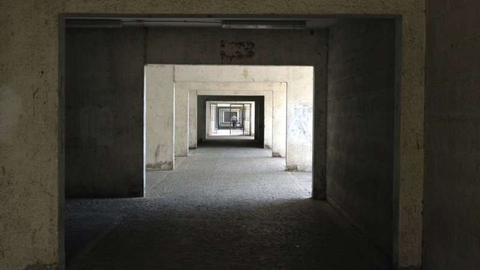 Corridoio interno lungo e rettilineo con una serie di aperture rettangolari allineate che creano una prospettiva ripetuta. Pareti in cemento grezzo e pavimento in pietra o graniglia, con luce naturale che filtra dalle aperture e una grata visibile in fondo.