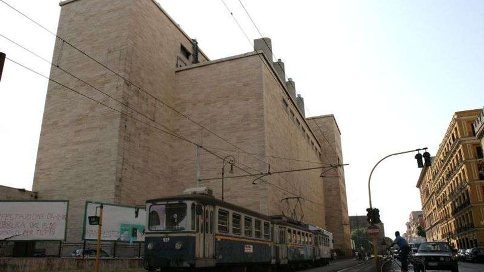 Grande edificio urbano in mattoni chiari con facciate quasi cieche e volumi squadrati, affacciato su una strada con binari del tram. In primo piano passa un tram sotto una rete di cavi elettrici; sul lato destro si vedono altri palazzi e un incrocio con semaforo.