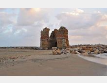 Rovine di una torre o fortificazione in pietra e mattoni sulla spiaggia sabbiosa, circondate da scogli e detriti rocciosi. Sullo sfondo si vede il mare con onde leggere e un cielo nuvoloso.