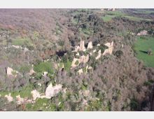 Veduta aerea di un complesso di rovine in pietra, con resti di torri e mura, immerso in una fitta area boschiva. Intorno si vedono pendii rocciosi e campi agricoli che si estendono nella campagna circostante.