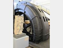 Interno di un museo tecnico con una grande turbina o generatore industriale in metallo scuro, con carter curvo forato e passerelle con ringhiere sullo sfondo. In primo piano c’è un piedistallo con un bassorilievo in pietra esposto vicino al macchinario.