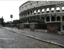 Veduta esterna del Colosseo con le sue arcate in pietra, ripreso da una strada in sampietrini e un marciapiede affiancato da muretti in mattoni e ringhiere metalliche. Sullo sfondo si vedono alberi e un cartello stradale.