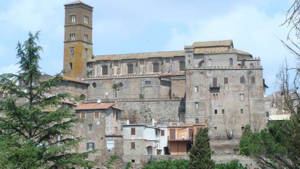 Complesso storico in pietra su un’altura, con un alto campanile quadrato e edifici fortificati con piccole finestre e tetti in coppi. In primo piano si vedono case in muratura e alberi, con vegetazione e cielo sullo sfondo.
