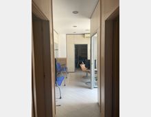 Corridoio interno di un ufficio con pavimento chiaro a listoni e pareti bianche, illuminato da faretti a soffitto. A sinistra ci sono sedute blu in fila, mentre in fondo si vede un’area reception con scrivania e computer, separata da una porta a vetri e con altri ambienti sullo sfondo.