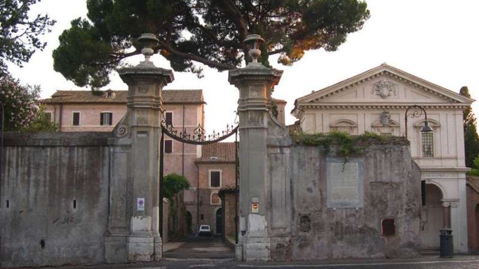 Ingresso monumentale con due pilastri in pietra e cancello in ferro battuto inseriti in un alto muro, con vialetto che conduce a edifici storici interni. Sullo sfondo si vedono grandi pini e una facciata in stile classico con frontone triangolare.