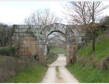 Antico arco in pietra inserito in un basso muro di blocchi squadrati, con tracce di muschio e usura. Un sentiero sterrato passa sotto l’arco e prosegue tra prati e alberi spogli in un paesaggio rurale.