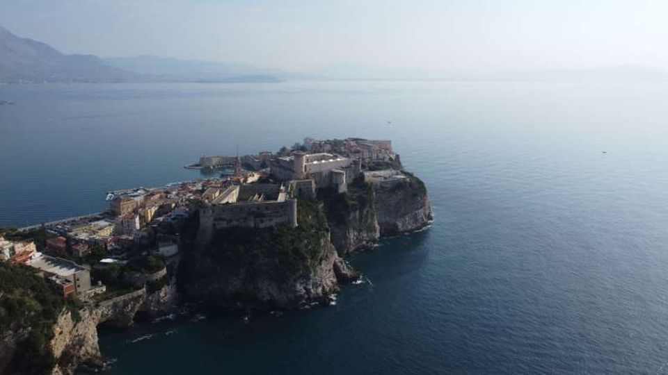 Veduta aerea di un borgo costiero fortificato con mura e bastioni in pietra, costruito su un promontorio roccioso a picco sul mare. Intorno si estende il mare aperto con costa e rilievi montuosi sfumati all’orizzonte.