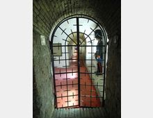 Ingresso ad arco in mattoni con cancello in ferro a grata, che conduce a un lungo corridoio sotterraneo illuminato, con pavimento rosso e muri bianchi. Lungo il passaggio si susseguono archi e nicchie, con pannelli informativi alle pareti.