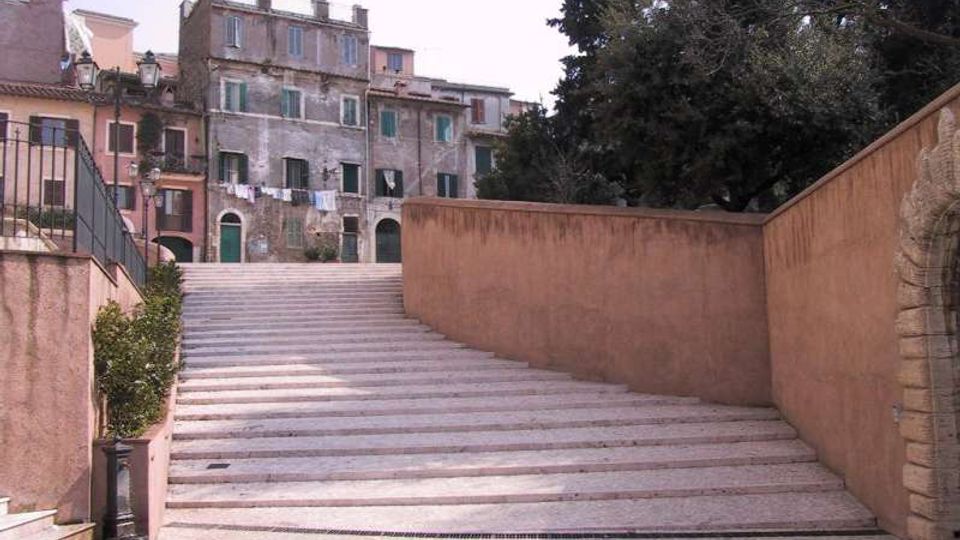 Ampia scalinata in pietra che sale tra muri intonacati color ocra, con lampione e ringhiera sul lato sinistro. In cima si affacciano edifici residenziali alti e vissuti con persiane e archi al piano terra, accanto a grandi alberi.