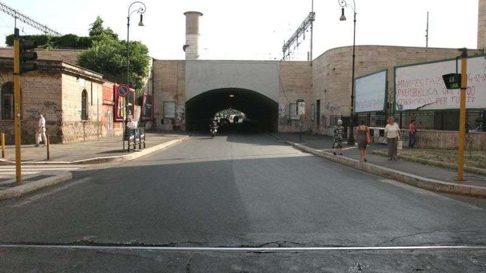 Strada urbana che conduce a un sottopasso ad arco in muratura, affiancato da edifici bassi con graffiti. Ai lati ci sono marciapiedi con lampioni, segnali stradali e cartelloni, con un incrocio in primo piano.
