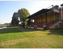 Tribuna coperta di un piccolo campo sportivo, con gradinate in muratura e struttura metallica a sostegno del tetto. Davanti si estende un prato verde, con alberi alti sullo sfondo e recinzione lungo il bordo del campo.