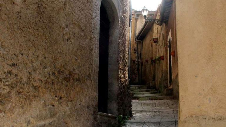 Vicolo stretto in un centro storico, con pavimentazione in pietra e alcuni gradini, fiancheggiato da alte pareti intonacate. Si vedono un portale ad arco, un piccolo balcone in ferro battuto e un lampione lungo il passaggio.