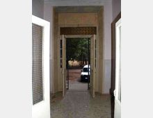 Ingresso interno con alte porte a battente e telaio in muratura chiara, che conduce all’esterno verso un cortile con alberi. Attraverso l’apertura si vede un vialetto in terra battuta e un veicolo parcheggiato.