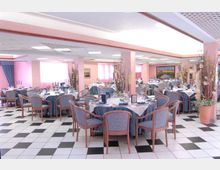 Ampia sala da pranzo interna con tavoli rotondi apparecchiati, sedie imbottite e decorazioni floreali alte. Pavimento a scacchiera bianco e nero, colonne rosa e soffitto con pannelli luminosi in un ambiente tipo ristorante o sala ricevimenti.
