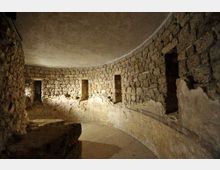 Interno di una struttura sotterranea in pietra con pareti curve in blocchi squadrati e soffitto a volta. Lungo il muro si aprono piccole nicchie rettangolari e il pavimento in terra segue l’andamento semicircolare dell’ambiente.