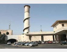 Parcheggio esterno di un complesso industriale in pietra chiara, con una alta torre cilindrica dotata di scala a spirale esterna. In primo piano sono visibili numerose auto parcheggiate, con un portico lungo e un’ampia apertura ad arco che conduce a un’area coperta.