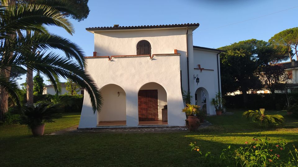 Edificio bianco in stile mediterraneo con portico ad archi e tetto in tegole, affacciato su un prato con palme e piante in vaso. Sul lato destro si vede una croce sulla facciata e un piccolo ingresso ad arco, circondati da alberi alti.