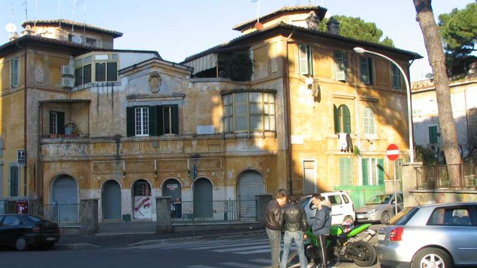 Edificio storico a più piani con facciata ocra e persiane verdi, con portici ad archi al piano terra e finestre ad arco al primo piano. Davanti si vede un incrocio con strisce pedonali, auto parcheggiate e un lampione, con alberi sullo sfondo.