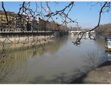 Veduta di un fiume urbano con alte banchine in muratura e palazzi storici lungo la riva sinistra, con alberi spogli in primo piano che incorniciano la scena. Sullo sfondo si vede un ponte e alcune imbarcazioni attraccate vicino alla riva destra.