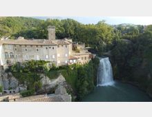 Veduta aerea di un edificio storico in pietra con torre quadrata e tetti in coppi, costruito su una rupe boscosa. Accanto all’edificio scende una cascata alta in un bacino d’acqua, circondato da alberi e colline.