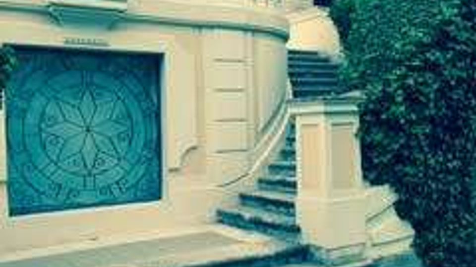 Scalinata in pietra che conduce a una grande porta decorata su una facciata chiara con balaustra e colonne, in un edificio residenziale. Ai lati ci sono siepi e alberi, con un prato verde in primo piano.