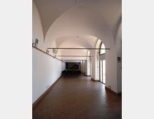 Interno di una sala rettangolare con soffitto a volte bianche e pavimento in mattoni, illuminata da grandi porte-finestre ad arco sul lato destro. In fondo si vedono file di sedie disposte come per una riunione o conferenza.