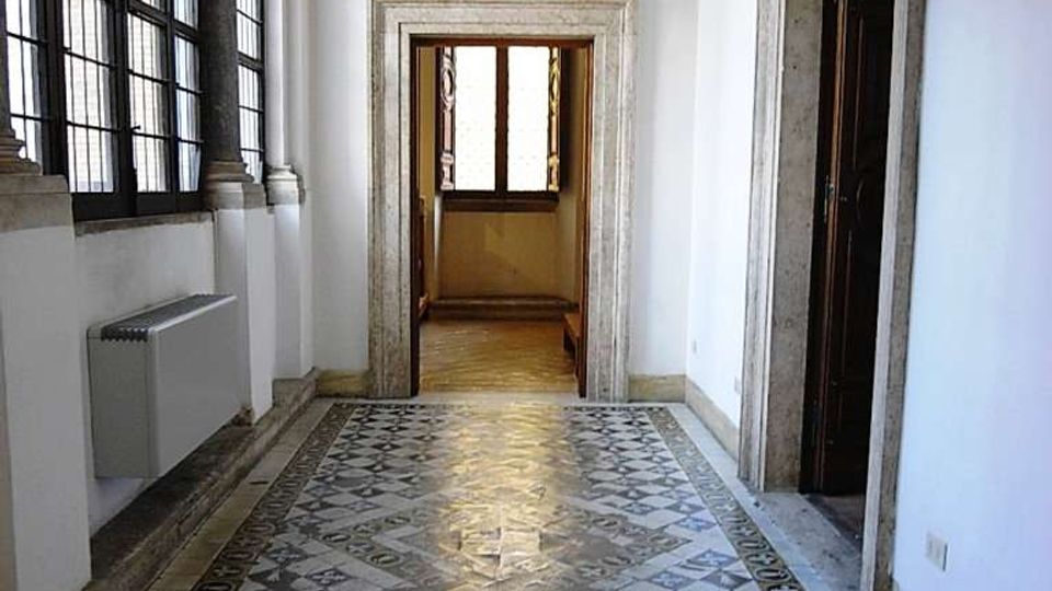 Corridoio interno di un edificio storico con pavimento in piastrelle decorative a motivi geometrici, grandi finestre a griglia sul lato sinistro e portali in pietra. Il soffitto è a volta con affreschi e cornici dorate, con un passaggio che conduce a una stanza illuminata in fondo.