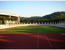 Vista di uno stadio all’aperto con pista di atletica rossa che circonda un campo in erba verde, con gradinate in cemento e una tribuna coperta sulla sinistra. Sullo sfondo si vedono colline boscose sotto un cielo sereno.