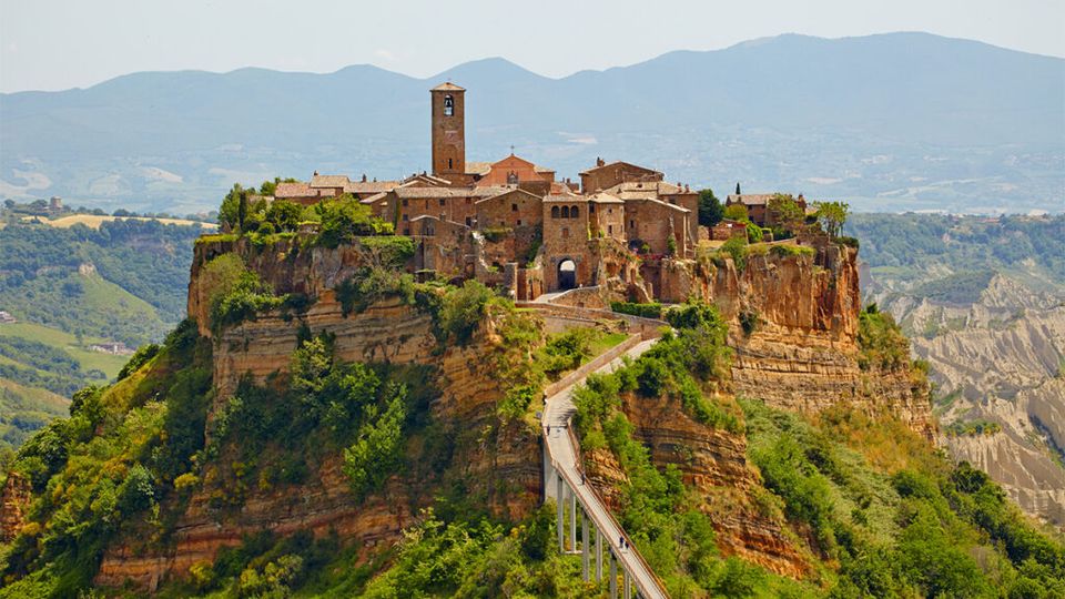 Borgo medievale in pietra arroccato su una rupe di tufo, con torre campanaria e case compatte affacciate su un ingresso ad arco. Un lungo ponte pedonale su piloni collega il paese al crinale, con colline e montagne sullo sfondo.