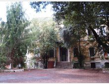 Ingresso di un edificio a due piani con facciata chiara, raggiunto da una breve scalinata centrale. L’area antistante è un cortile in terra battuta circondato da alberi alti e vegetazione fitta che ombreggia la struttura.