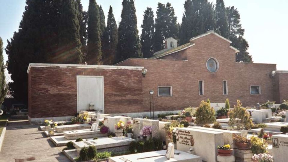 Cimitero con tombe in marmo e vasi di fiori su un vialetto pavimentato, davanti a un edificio in mattoni rossi con piccola finestra rotonda e porta laterale. Sullo sfondo si vedono alti cipressi che circondano l’area.