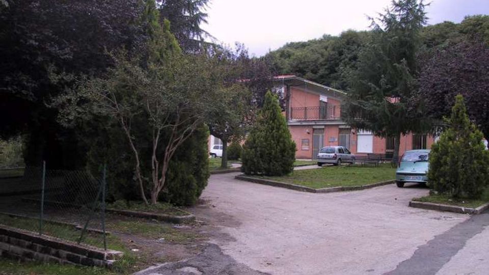 Strada asfaltata interna con parcheggio e aiuole alberate, circondata da alberi alti e vegetazione fitta. Sullo sfondo si vede un edificio basso in mattoni rosa con balconi e tetto spiovente, con alcune auto parcheggiate.
