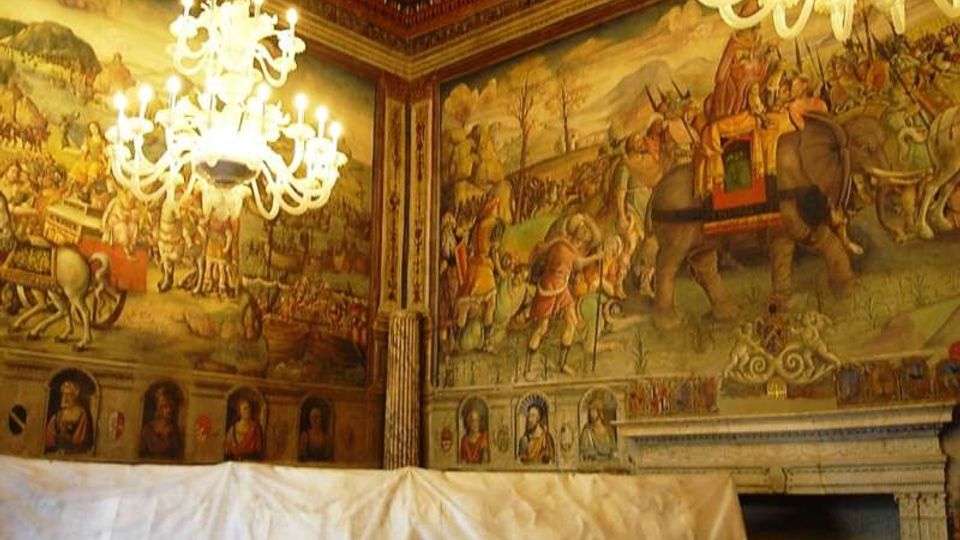 Salone interno con pareti affrescate raffiguranti scene storiche e figure, soffitto a cassettoni decorato e grandi lampadari in vetro sospesi. In basso si vede un ampio telo bianco che copre parte della sala e una grande cornice architettonica simile a un camino.