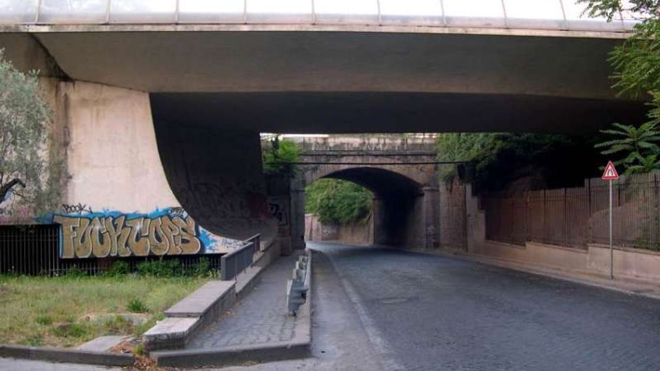Sottopasso stradale urbano in cemento con carreggiata in sampietrini e marciapiede laterale, sotto un grande viadotto. Sulle pareti sono presenti graffiti e, in fondo, si vede un secondo arco in muratura con vegetazione ai lati.