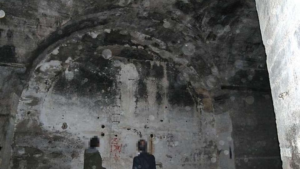 Interno di un ambiente sotterraneo in cemento, simile a un bunker, con soffitto a volta e pareti grezze macchiate e scrostate. Sulla parete di fondo si notano una nicchia ad arco, fori e piccoli graffiti, con illuminazione fioca e ombre marcate.