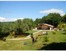 Casa di campagna a un piano con tetto in legno e ampia veranda, situata su un prato in pendenza e collegata da un vialetto chiaro. Intorno ci sono alberi e vegetazione, con colline boscose sullo sfondo sotto un cielo azzurro.