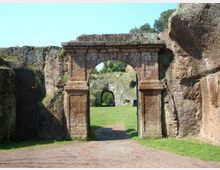 Portale in pietra con arco centrale incastonato tra alte pareti rocciose, che introduce a un prato con un sentiero. In fondo si vede un secondo arco più piccolo ricavato nella roccia.