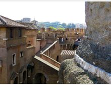 Cortile e passaggi sopraelevati di una fortificazione storica in mattoni, con tetti in tegole e mura merlate. Un camminamento con parapetti e scalinate attraversa la struttura, con la città e colline alberate sullo sfondo.