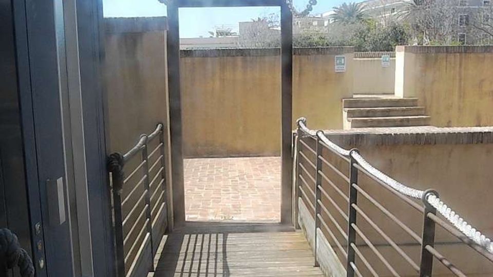 Passerella in legno con corrimano metallici e catene laterali, che conduce a un’uscita rettangolare verso un cortile pavimentato in mattoni. Sullo sfondo ci sono muretti in cemento, una breve scalinata e edifici lontani sotto un cielo sereno.