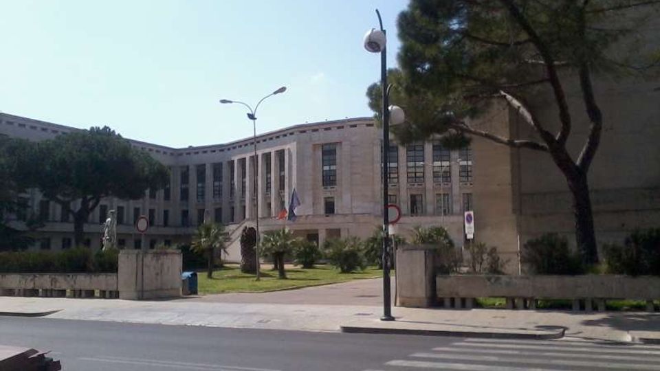Grande edificio istituzionale in pietra chiara con facciata curva, alte finestre verticali e ingresso centrale con bandiere. Davanti si vede una strada con strisce pedonali, lampioni e un’area verde con alberi e aiuole.