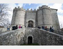 Castello in pietra con due grandi torri cilindriche merlate e portale centrale, visto dal ponte in muratura che conduce all’ingresso. Intorno si notano mura in pietra, un fossato sottostante e alberi spogli sullo sfondo sotto un cielo parzialmente nuvoloso.