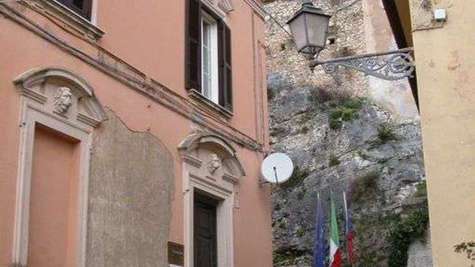 Scalinata in pietra in un vicolo stretto tra edifici storici intonacati, con finestre e portale decorato. In fondo si vede una parete rocciosa con una lanterna a muro e bandiere vicino ai gradini, sotto cavi sospesi.